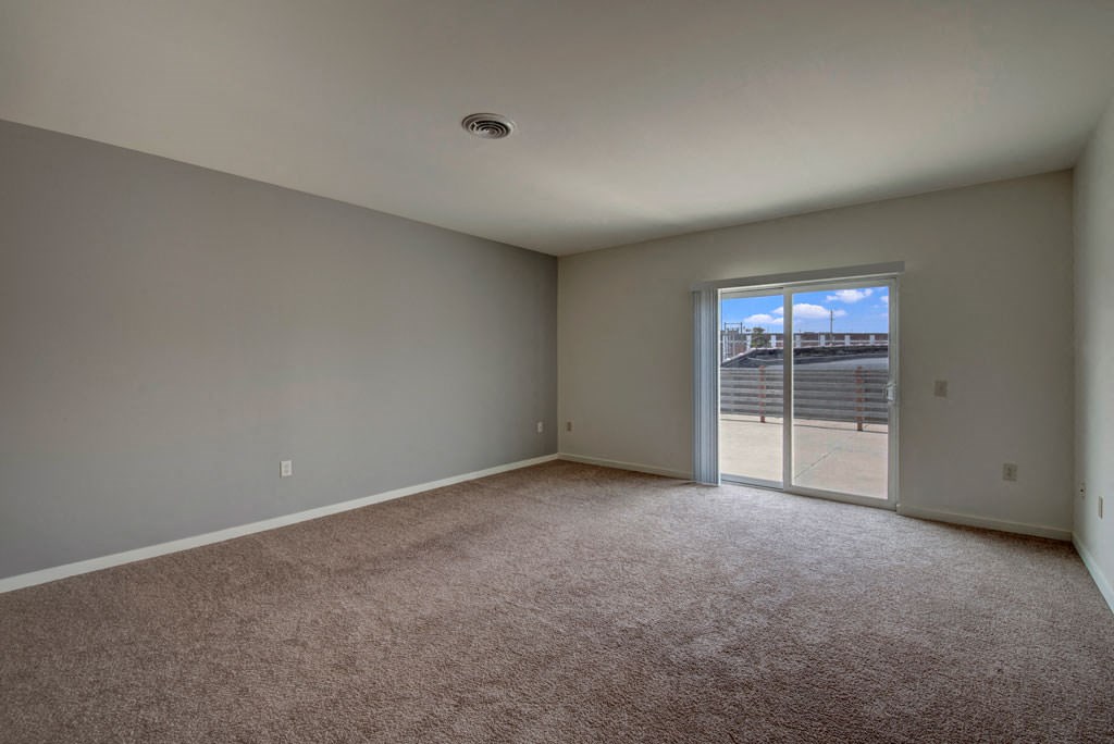 An empty room at Latitude Apartments with beige carpet and light gray walls features a large sliding glass door leading to a patio with a view of a clear blue sky.