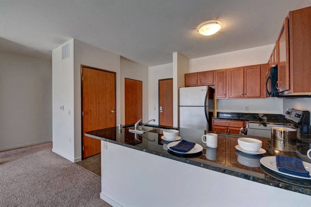Modern kitchen at the Latitude Apartments with wood cabinets, stainless steel appliances, and a black countertop bar set with plates, bowls, and white mugs, creating an inviting atmosphere.