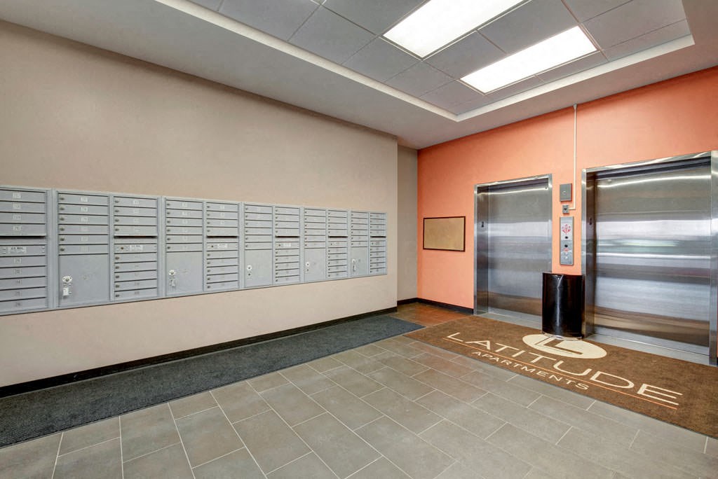 The Latitude Apartment lobby with gray mailboxes on a beige wall, two closed elevators on an orange wall, and a brown mat reading "Latitude Apartments" on tiled flooring.