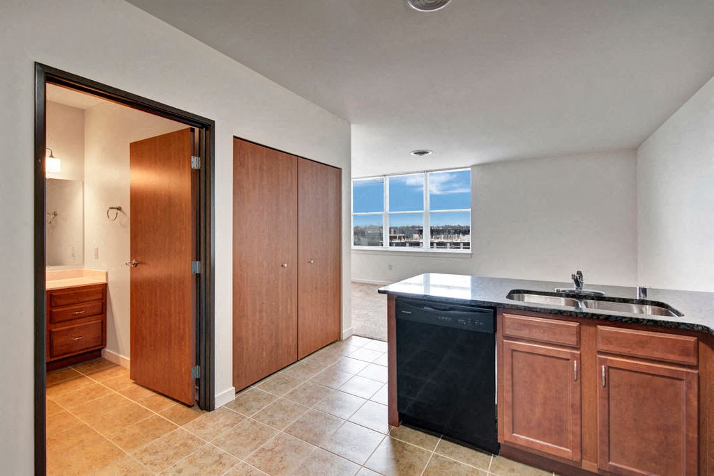 Modern apartment interior with open kitchen, featuring wooden cabinets, black counters, and tile floor. Bright window view and a closed bathroom door. The Latitude Apartments.