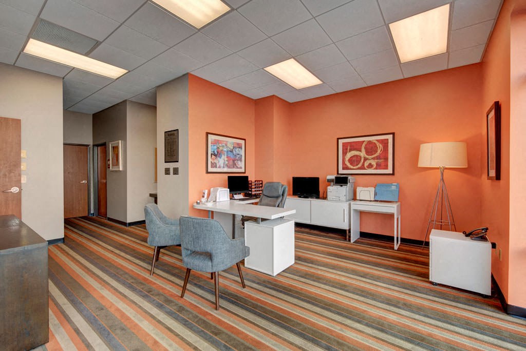 A modern business center with peach walls and striped carpet. Features a white desk with a computer and gray chairs. Artwork and a tall lamp add warmth. The Latitude Apartments.