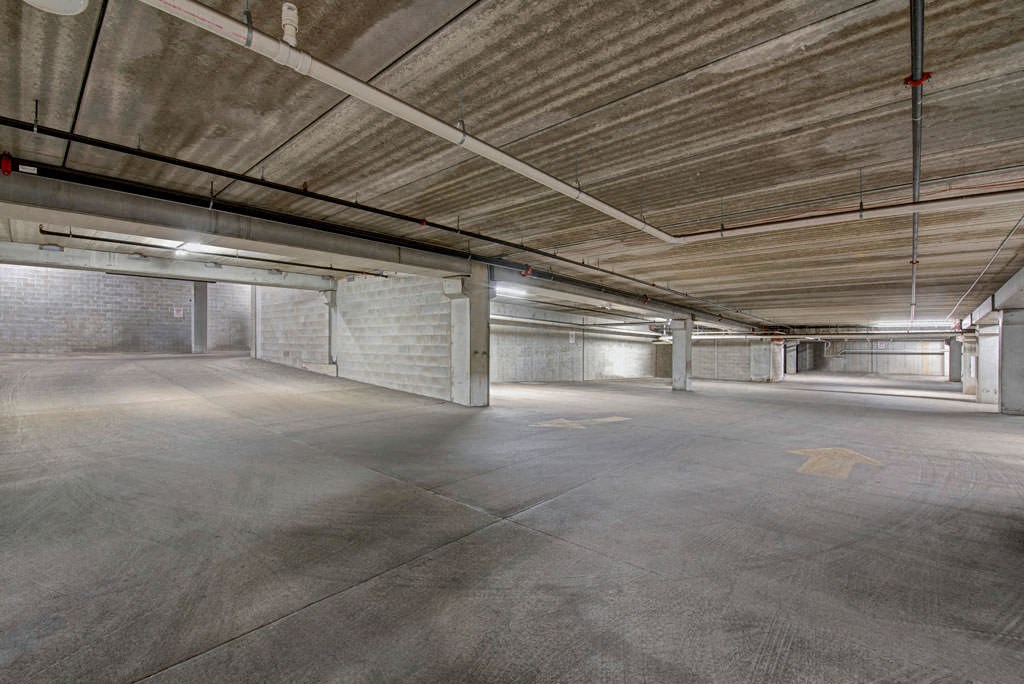 Empty underground parking garage at the Latitude Apartments with concrete walls, ceiling, and floor. Bright fluorescent lights illuminate the space, creating a stark, industrial feel. Yellow arrows mark directions on the floor.