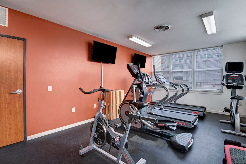 A gym at Latitude Apartments with a stationary bike, treadmills, and an elliptical facing two wall-mounted TVs. Large windows let in natural light, creating a clean, energizing atmosphere.