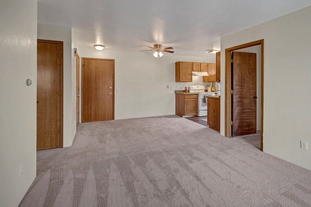 Spacious living area here at Legacy West Apartments with beige carpet and neutral walls, connected to a kitchen with wooden cabinets and white appliances. Ceiling fan above adds warmth.