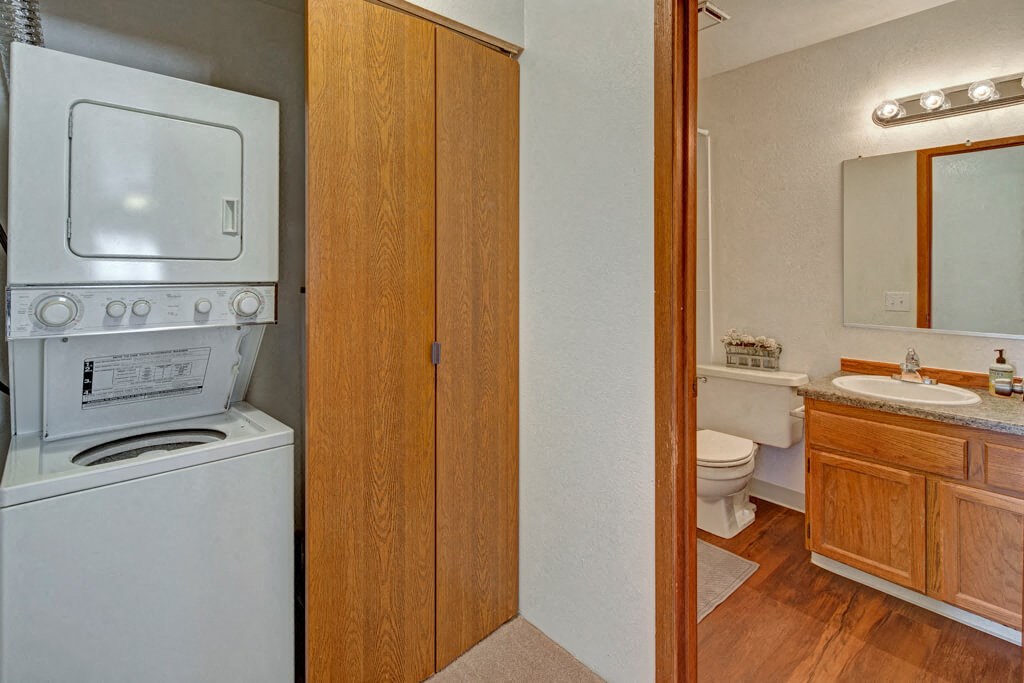Laundry area here at Legacy West Apartments with a stacked washer and dryer next to wooden cabinets. Adjacent bathroom with sink, mirror, toilet, and warm lighting; cozy atmosphere.