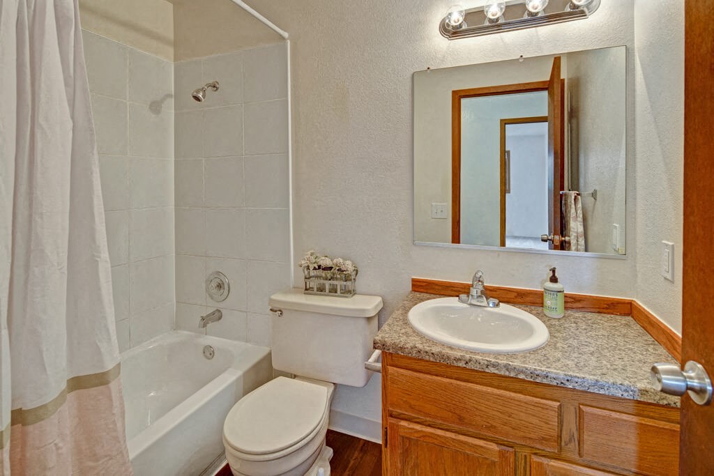 Small bathroom here at Legacy West Apartments with white tiled shower-tub combo, beige curtain, wooden vanity, granite counter, oval sink, mirror, and chrome light fixture.