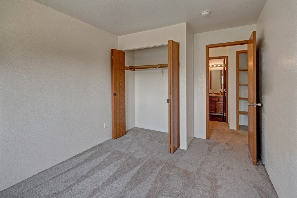 Empty bedroom here at Legacy West Apartments with light gray carpet, open wooden closet doors, and an adjoining bathroom door. Neutral walls and a tidy ambiance.