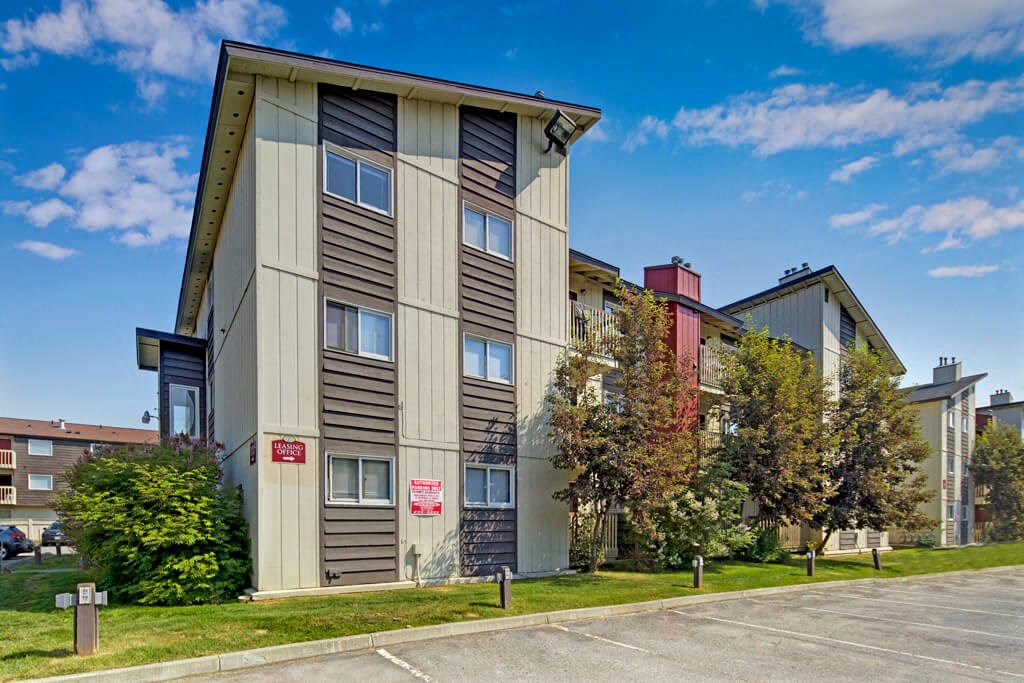 Three-story Legacy West apartment building with tan and brown siding, surrounded by trees and grass. Clear blue sky, empty parking lot in the foreground.