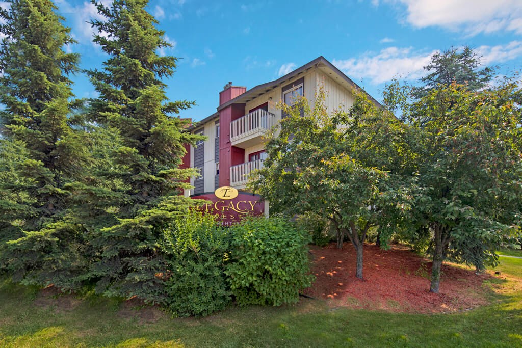 Legacy West Apartment building surrounded by lush trees and greenery under a blue sky. A sign reads "Legacy" in the foreground, creating a peaceful ambiance.