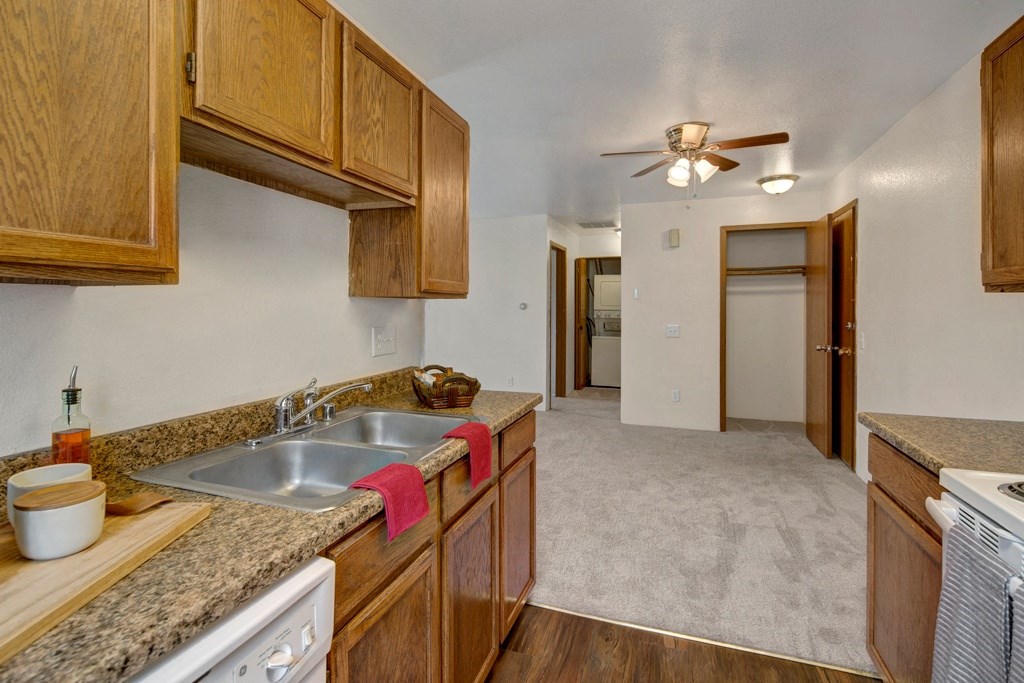 Kitchen here at Legacy West Apartments with wood cabinets and granite countertops, double sink with red towel, leading to carpeted hall. Ceiling fan and open closet visible. Cozy ambiance.