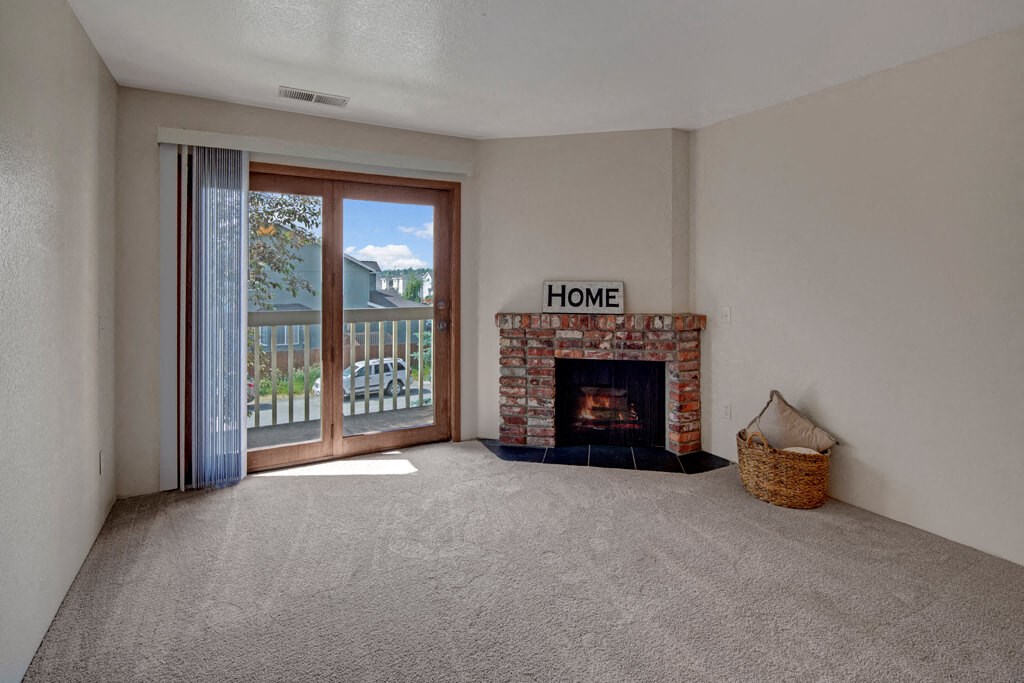 Empty living room here at Legacy West Apartments with a cozy brick fireplace, sign reading "Home," and a wicker basket on plush carpet. Sunlit balcony doors reveal trees outside.