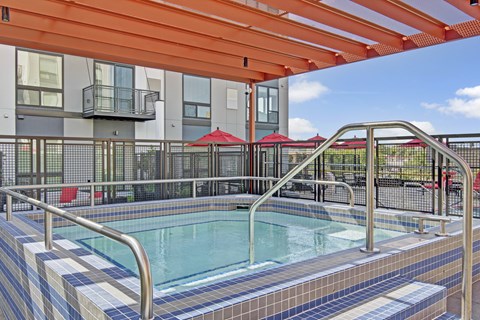 A modern outdoor spa here at 4800 Excelsior featuring a tiled hot tub with built-in steps and stainless handrails beneath a bold orange pergola. The fenced courtyard includes contemporary apartment façades, red patio umbrellas, and seating areas, creating a vibrant, inviting space for relaxation and socializing.