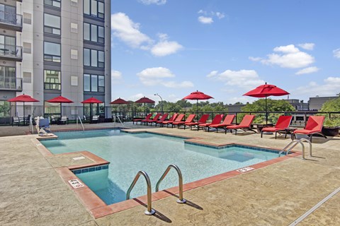 A resort-style outdoor pool here at 4800 Excelsior showcasing a rectangular swimming pool with clear blue water, stainless handrails, and a spacious concrete deck. Red lounge chairs and matching umbrellas line the poolside, with modern apartment buildings and treetop views creating an open, relaxing atmosphere.