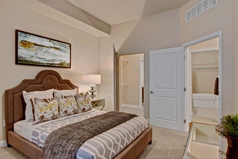 A serene bedroom here at 4800 Excelsior featuring a plush upholstered bed with patterned bedding, layered accent pillows, and a textured throw. Soft carpeting and neutral walls create a calming atmosphere, complemented by a bedside lamp, framed artwork, and a mirrored nightstand, with easy access to a walk-in closet and adjoining bathroom.