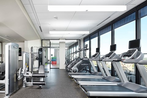 A bright fitness center here at 4Marq featuring rows of modern treadmills and stair machines positioned along expansive floor-to-ceiling windows with city views. Strength training equipment, open walkways, and sleek lighting create a spacious, clean, and motivating environment designed for a full range of workout routines.