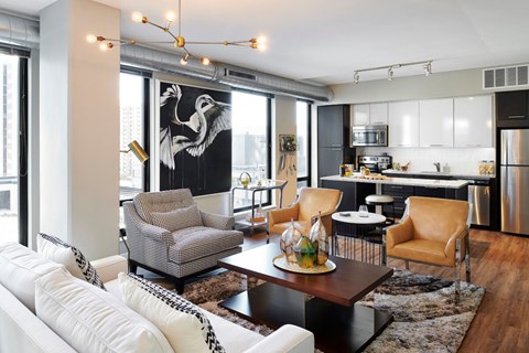 A modern open-concept community lounge here at 4Marq showcasing a stylish seating area with a white sofa, patterned armchair, and leather accent chairs centered around a low coffee table. Floor-to-ceiling windows, exposed ductwork, contemporary lighting, and a sleek kitchen backdrop create a bright, urban atmosphere.