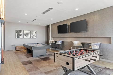 A modern game lounge here at 4Marq featuring a foosball table in the foreground and a billiards table set on patterned flooring. Two wall-mounted flat-screen televisions sit above a sleek counter with bar-height seating, while neutral walls, framed abstract artwork, and recessed lighting create a relaxed and social atmosphere.