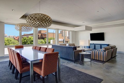 A contemporary community lounge here at 7West featuring a long dining table with upholstered chairs, modern pendant lighting, and a cozy seating area with sofas and armchairs. Large windows provide city views and natural light, while a mounted TV, accent tables, and clean finishes create a welcoming space for gatherings or relaxation.