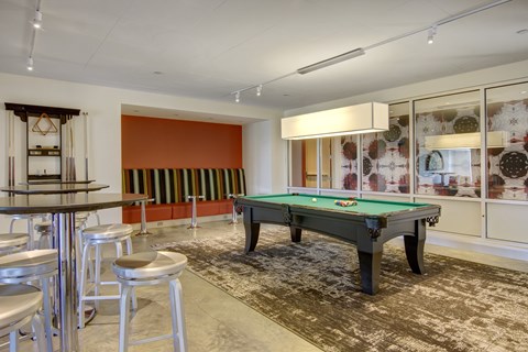 A lively game lounge here at 7West featuring a pool table with green felt centered on a patterned area rug. High-top tables with stools sit nearby, along with built-in bench seating against a bold accent wall. Glass panels, modern lighting, and recreational cues create a social space for games and casual gatherings.