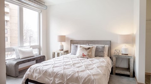 A serene bedroom here at 7West featuring a neatly made bed with crisp white bedding and soft accent pillows, flanked by matching bedside tables with modern lamps. A large window with a built-in bench seat allows natural light to fill the space, while clean white walls, exposed ductwork, and polished concrete flooring create a calm, contemporary atmosphere.