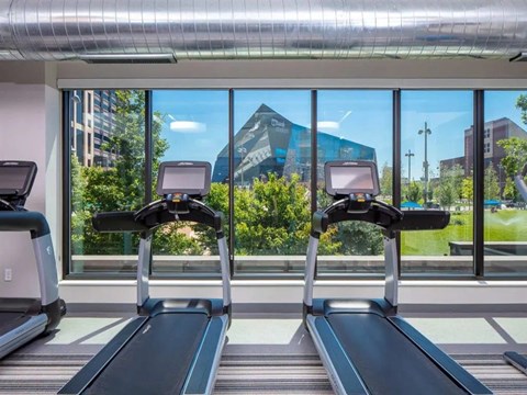 A modern fitness center here at Edition featuring treadmills positioned in front of floor-to-ceiling windows with views of landscaped greenery and nearby city buildings. Exposed ductwork, bright natural light, and clean-lined equipment create an open, energizing workout space designed for daily fitness routines.