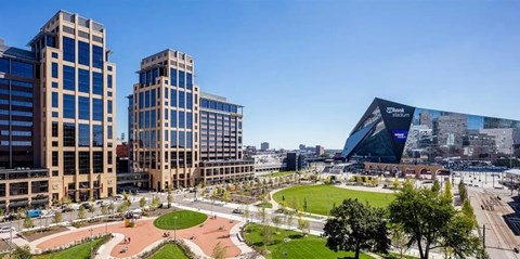 A panoramic cityscape view here at Edition overlooking a landscaped urban park with walking paths, open green lawns, and surrounding modern high-rise buildings. The scene features a prominent glass-fronted stadium nearby, tree-lined streets, and expansive blue skies that highlight the vibrant downtown setting.