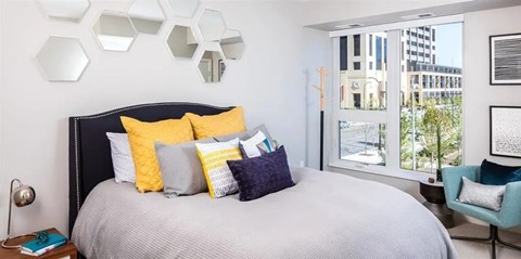 A stylish bedroom here at Edition showcasing a modern bed with layered neutral and yellow accent pillows, a textured upholstered headboard, and decorative wall mirrors above. Large windows bring in natural light and offer city views, while coordinated furnishings and soft tones create a comfortable, polished retreat.