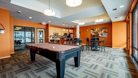 A lively community lounge here at Gallery Flats featuring a full-size pool table centered on patterned carpet flooring. High-top tables with seating, warm accent walls, and modern lighting surround the space, while large windows and glass doors bring in natural light and connect the lounge to nearby community areas.