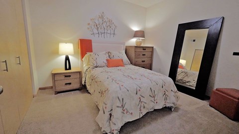 A cozy bedroom here at Gallery Flats with soft beige carpet, neutral walls, and a comfortable bed dressed in patterned bedding with accent pillows. Matching bedside tables with lamps provide warm lighting, while a tall dresser and a full-length mirror add function and style to the inviting space.