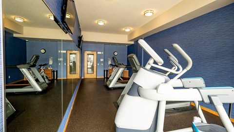 A modern fitness center here at Gallery Flats featuring cardio machines arranged along mirrored walls that reflect the space and lighting. Blue accent walls, dark rubber flooring, and ceiling-mounted lights create a clean, energized environment, while a wall-mounted television and glass doors add convenience and visibility.