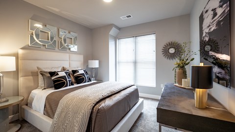A cozy bedroom here at Gatsby with plush carpet flooring, soft neutral walls, and a padded headboard bed dressed in layered bedding and decorative pillows. A window with blinds brings in natural light, while bedside tables with lamps, wall art, and a dresser create a comfortable, thoughtfully styled retreat.
