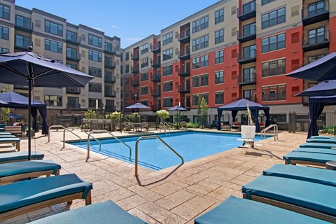 A resort-style pool courtyard here at Gatsby featuring a rectangular swimming pool surrounded by cushioned lounge chairs, shaded umbrellas, and private cabana seating. Modern apartment buildings with balconies frame the space, while clean stone decking and landscaped greenery create an inviting outdoor area for relaxation and socializing.