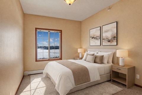 A cozy bedroom here at Grand River Estates featuring soft neutral walls, plush carpet flooring, and a comfortable bed with layered white bedding and accent pillows. The room includes matching nightstands with lamps, framed wall art above the headboard, and a window that brings in natural light and outdoor views, creating a calm and inviting sleeping space.