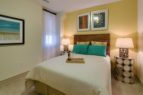 A cozy bedroom here at Inspire featuring a woven headboard bed with soft neutral bedding, layered accent pillows, and matching bedside tables topped with modern lamps. Warm wall tones, framed artwork, sheer window treatments, and plush carpeting create a relaxing retreat with inviting, thoughtfully styled details.