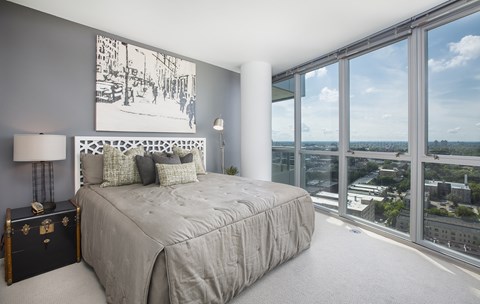 A sunlit bedroom here at LPM Apartments featuring a comfortable bed dressed in soft neutral bedding and layered accent pillows, set against a gray accent wall with modern artwork, while expansive floor-to-ceiling windows wrap the space with sweeping city views, filling the room with natural light and creating an airy, serene retreat.