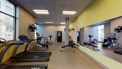 A modern fitness center here at Lofts at Farmers Market featuring treadmills and cardio machines lined along large windows, mirrored walls that reflect the workout space, a weight bench and exercise equipment for strength training, neutral flooring, light-colored walls, ceiling lighting, and an open layout designed for convenient and comfortable workouts.