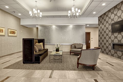 A stylish resident lounge living room here at Lofts at Farmers Market featuring elegant chandeliers, patterned accent walls, a sleek fireplace, and comfortable seating arranged around a glass coffee table, with tiled flooring and soft area rugs creating a refined yet inviting space for relaxing or socializing.