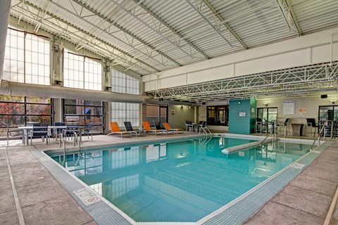 A spacious indoor pool here at Marbella on Dean featuring a large rectangular swimming pool with clear blue water, stainless steel handrails, surrounding concrete deck, and lounge seating with bright accent chairs, set beneath a high industrial-style ceiling with exposed beams and large windows that bring in natural light.
