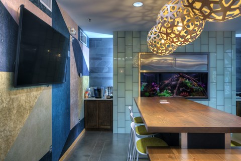 A modern community gathering space here at Marbella on Dean featuring a long wooden table with contemporary seating beneath sculptural pendant lighting, accented by textured walls, a large wall-mounted television, and a built-in aquarium centerpiece, with polished concrete floors and adjacent refreshment cabinetry creating a stylish and functional social setting.