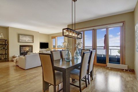 A bright open dining and living area here at The Mist featuring light wood-style flooring, a rectangular dining table with upholstered chairs, and a modern pendant light overhead. The space flows into a cozy living room with a fireplace and seating, while large glass doors and windows showcase scenic water views and lead to a private balcony.