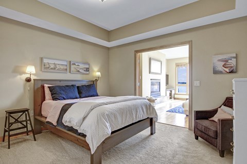 A serene bedroom here at The Mist featuring a spacious layout with a wood-framed bed dressed in soft neutral bedding, matching wall-mounted lamps, and framed coastal artwork above the headboard. Plush carpeting adds comfort underfoot, while an open doorway leads to a bright adjoining living area with large windows that bring in abundant natural light and scenic views.