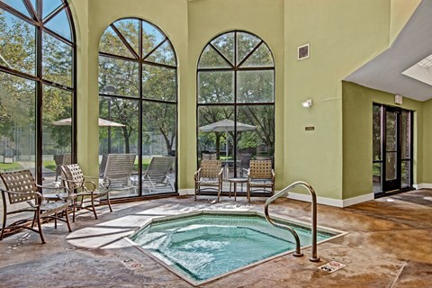 A serene indoor hot tub area here at Plymouth Square featuring a square spa with gently bubbling water, stainless handrails, and a tiled deck surround. Tall arched windows bring in abundant natural light and views of outdoor seating, while nearby lounge chairs and neutral finishes create a calm, resort-inspired atmosphere.