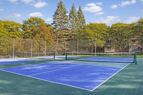 A scenic outdoor tennis court here at Plymouth Square featuring blue and green playing surfaces, a central net, and surrounding fencing, set against a backdrop of mature trees and nearby apartment buildings. The open layout and clear markings create an inviting space for residents to enjoy casual matches or practice in a peaceful, park-like setting.