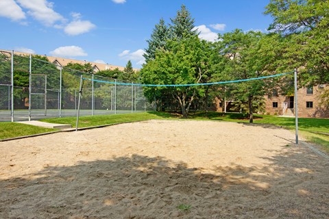 A spacious outdoor sand volleyball court here at Plymouth Square featuring a level sandy playing surface with a central net, bordered by green lawn, mature trees, and nearby apartment buildings, creating a relaxed, park-like setting where residents can enjoy casual games or outdoor recreation on sunny days.