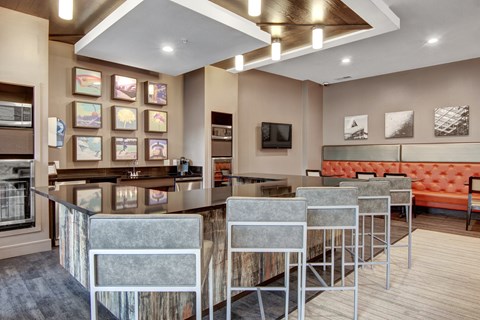 A contemporary resident lounge here at Residence at the COR showcasing a bar-height island with cushioned stools and sleek countertops, complemented by warm wood accents, an orange tufted banquette, modern wall art, and layered recessed and pendant lighting, all within an open, inviting layout designed for socializing, entertaining, and everyday relaxation.