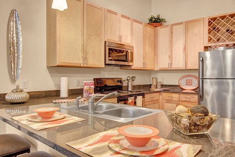 A contemporary kitchen here at Residence at the COR showcasing light wood cabinetry, sleek countertops with an integrated double sink, stainless-style appliances, and a built-in microwave, complemented by a breakfast bar set with place settings, decorative accents, and warm pendant lighting that creates a functional and inviting cooking space.
