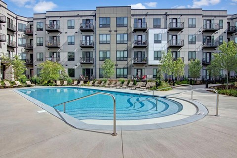 A resort-style outdoor pool here at Residence at the COR featuring a curved entry with handrails and shallow steps, surrounded by a wide concrete deck lined with lounge chairs, while a modern apartment building with private balconies and landscaped greenery creates a polished, inviting backdrop for relaxation and social time.