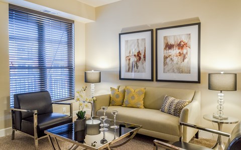 A warm and inviting living room here at Siena featuring a comfortable sofa accented with patterned throw pillows, a sleek glass coffee table, and modern table lamps that provide soft lighting, complemented by contemporary wall art, a cozy accent chair, and a large window with blinds that brings in natural light and creates a relaxed, welcoming atmosphere.