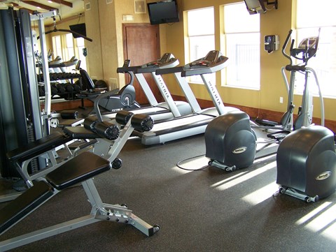 A well-equipped fitness center here at Stoneleigh at the Reserve featuring treadmills, elliptical machines, and strength-training equipment arranged on rubber flooring, with large windows bringing in natural light, wall-mounted televisions, mirrors, and warm neutral walls creating a bright and functional workout space.