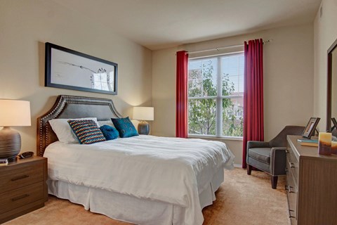 A cozy bedroom here at Stoneleigh at the Reserve featuring a comfortable bed with crisp white bedding and colorful accent pillows, complemented by matching nightstands with lamps, soft carpeted flooring, and neutral walls, while a large window with curtains brings in natural light and creates a calm, inviting retreat for rest and relaxation.